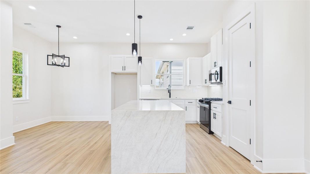 Modern, bright kitchen with white cabinetry, a sleek marble like island, wood flooring, and contemporary pendant lighting. The open layout and large window enhance the airy feel.