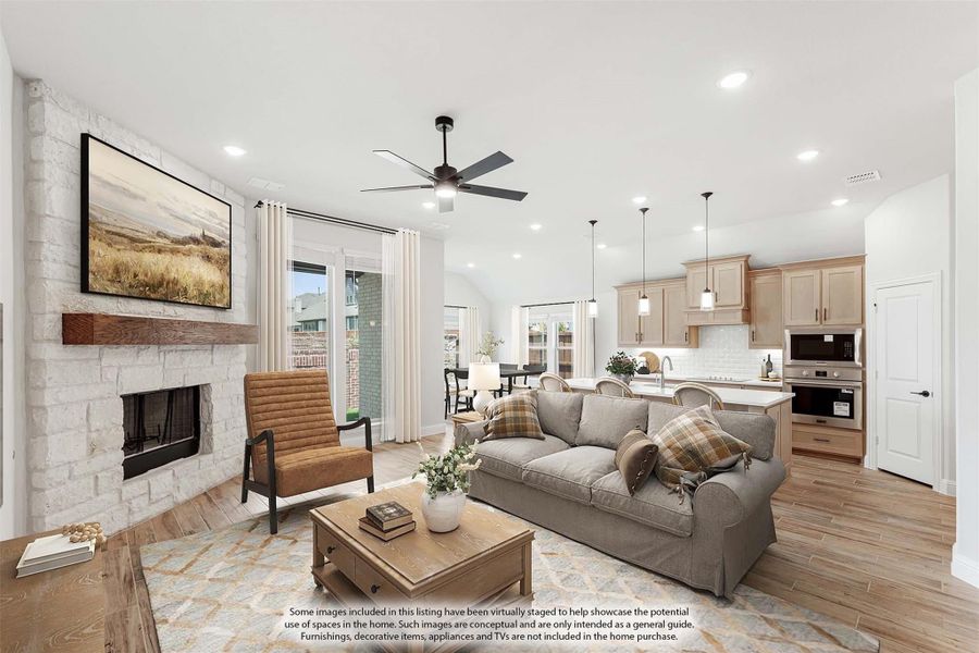 Open-concept living room with stone fireplace, ceiling fan, and kitchen visible in background on wood-look floors.
