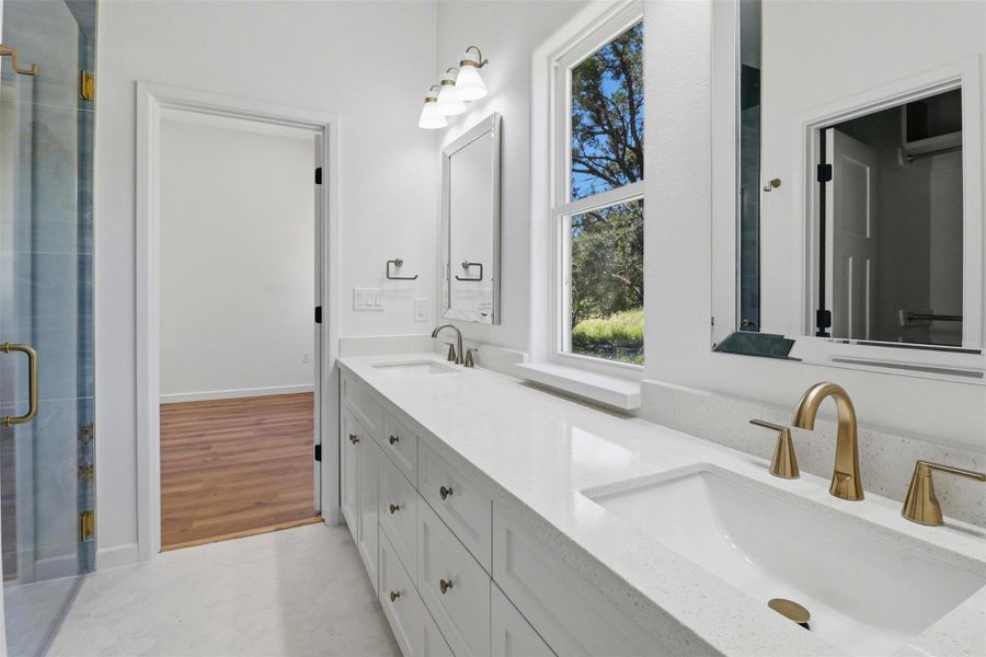 Dual vanity with white cabinetry, light-colored countertops, and gold-finish fixtures Dual vanity with white cabinetry, light-colored countertops, and gold-finish fixtures