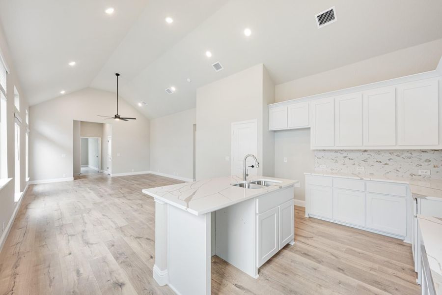 Open kitchen with white cabinets, marble island, and light wood floors opening to living area with vaulted ceiling