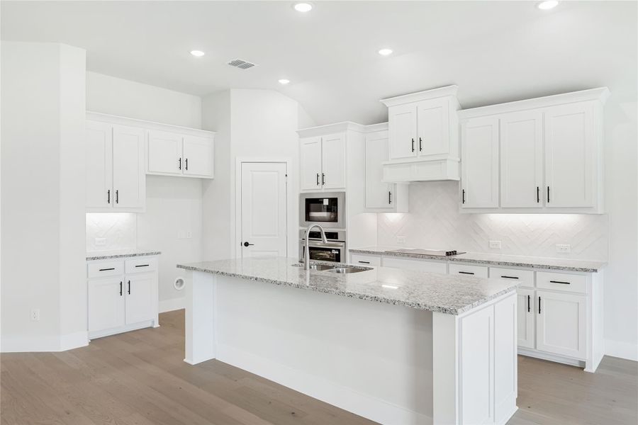 White kitchen with large granite island, white shaker cabinets, stainless steel appliances, and light wood floors