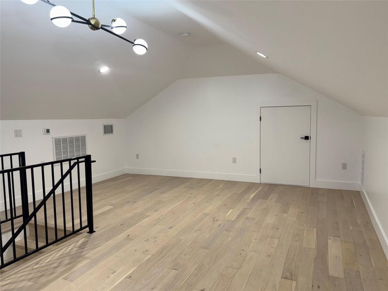 Spacious loft area featuring light wood-finish flooring, a modern chandelier, recessed lighting, a black metal railing, and a white interior door