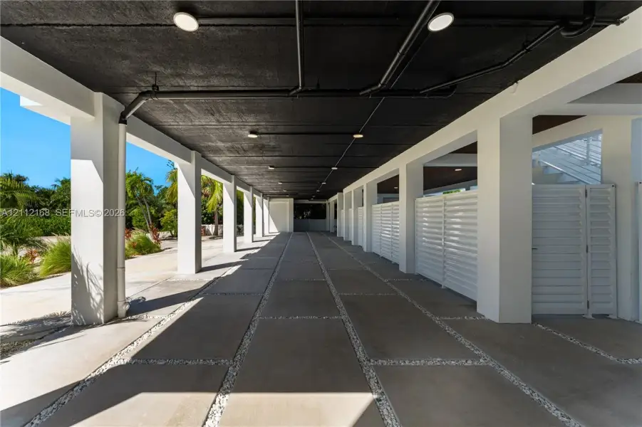 Covered ground-level parking area featuring concrete pavers, white louvered privacy screens, recessed lighting, and exposed ceiling structure Covered ground-level parking area featuring concrete pavers, white louvered privacy screens, recessed lighting, and exposed ceiling structure