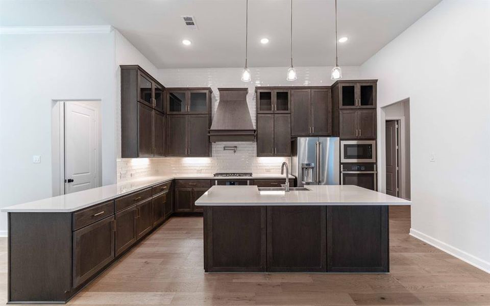 Open-concept kitchen featuring dark wood-finish cabinetry, light countertops, a central island with a sink, stainless steel appliances, and wood-finish flooring Open-concept kitchen featuring dark wood-finish cabinetry, light countertops, a central island with a sink, stainless steel appliances, and wood-finish flooring