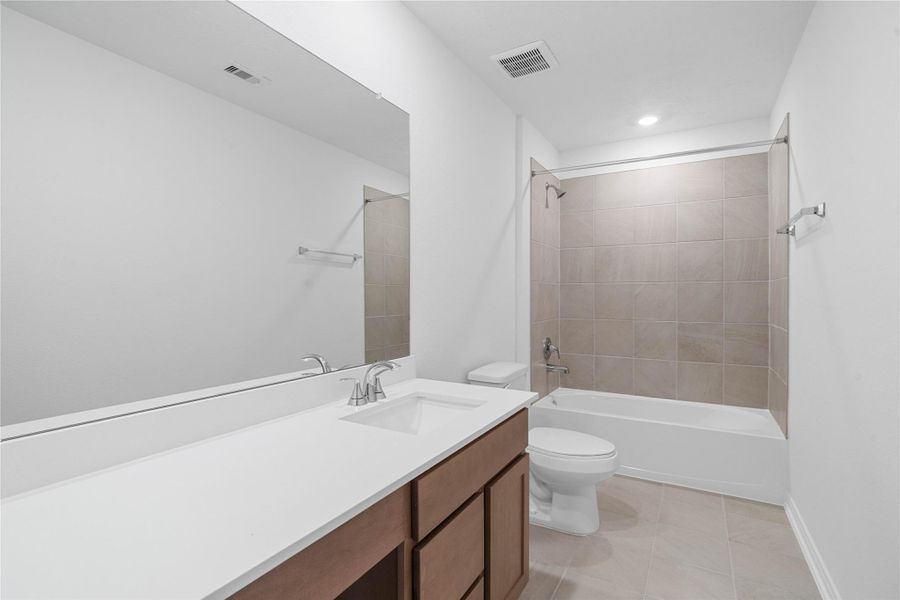 The secondary bathroom showcases tile flooring, a bath/shower combo with Daltile tile surround, a vanity with knee space and stunning light countertop, a mirror, sleek fixtures, and contemporary finishes throughout.
