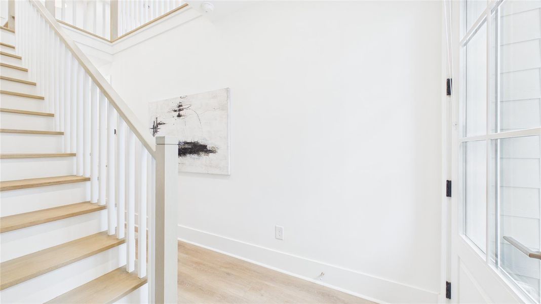 Bright and airy entryway with light wood flooring and stairs, white walls, and a large windowed door. The space features contemporary decor and a clean, modern aesthetic.