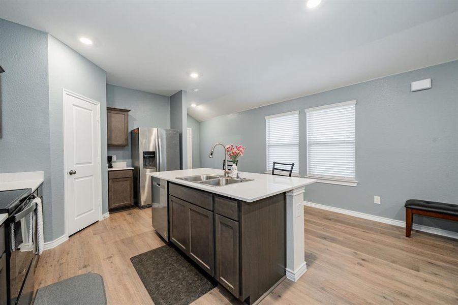Kitchen with dark brown cabinets, light wood-type flooring, an island with sink, appliances with stainless steel finishes, and vaulted ceiling Kitchen with dark brown cabinets, light wood-type flooring, an island with sink, appliances with stainless steel finishes, and vaulted ceiling