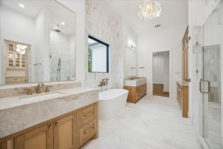 The acrylic soaking tub sits against natural stone with generous light, creating a serene spa-like setting. Dual vanities offer storage, supported by a high-efficiency tankless water heater for continuous hot water.