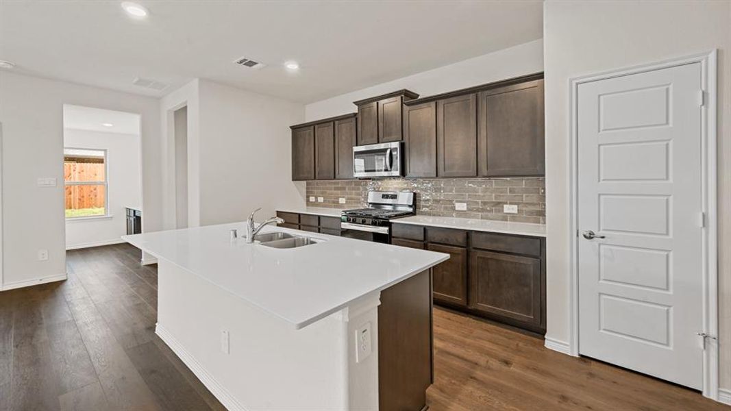Kitchen featuring a large island with a double basin sink, dark wood-finish cabinetry, light-toned countertops, and a brick-patterned backsplash Kitchen featuring a large island with a double basin sink, dark wood-finish cabinetry, light-toned countertops, and a brick-patterned backsplash