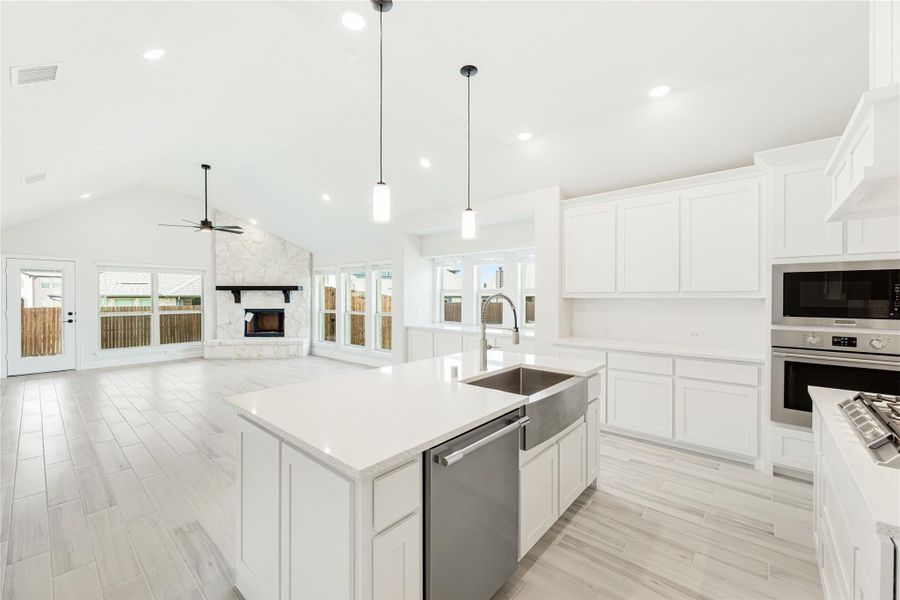 Open kitchen with white cabinets, large island, stainless appliances, and vaulted ceiling open to living room with stone fireplace.
