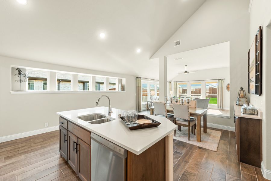 Kitchen island with double sink and dishwasher, open to dining room with vaulted ceiling and wood-look floors