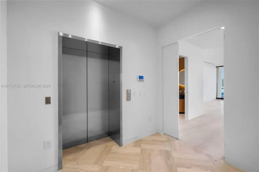 Stainless steel elevator with brushed metal finish, herringbone wood-finish flooring, and recessed ceiling lighting