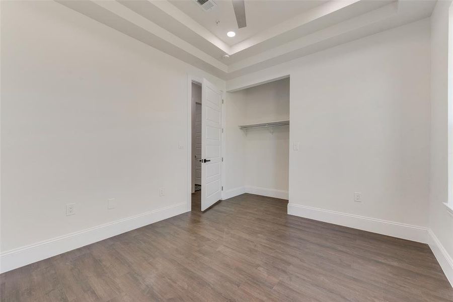 Room featuring wood-finish flooring, crisp white walls, a tray ceiling with recessed lighting, and a built-in closet with shelving Room featuring wood-finish flooring, crisp white walls, a tray ceiling with recessed lighting, and a built-in closet with shelving