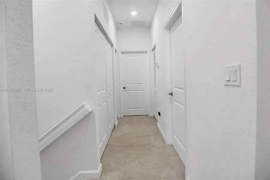 Hallway featuring neutral wall-to-wall carpeting, three paneled interior doors, a double-door closet, and recessed ceiling lighting Hallway featuring neutral wall-to-wall carpeting, three paneled interior doors, a double-door closet, and recessed ceiling lighting