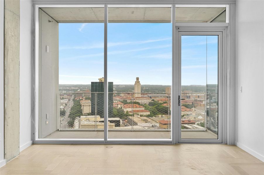 Living room with large, floor-to-ceiling glass doors leading to a balcony. The view overlooks a cityscape with prominent buildings, including the University of Texas. The room has light-colored wood flooring and a clean, modern aesthetic with a minimalist design. The balcony is enclosed with a transparent safety railing, offering an unobstructed view of the skyline and surrounding area under a clear, blue sky.