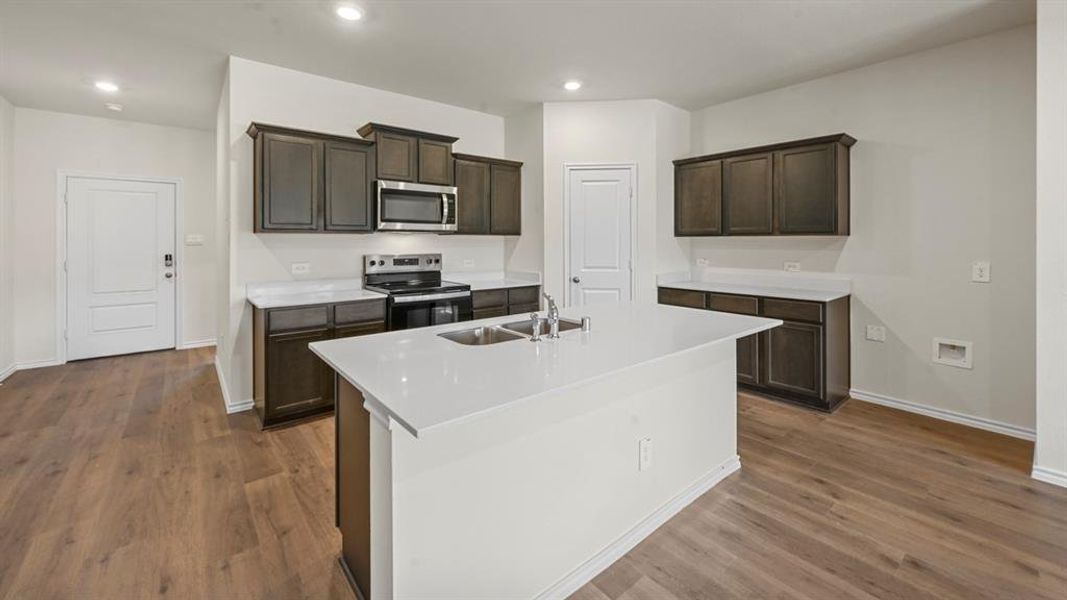 Spacious kitchen featuring an island with an undermount sink, dark wood-finish cabinetry, white countertops, stainless steel appliances, and wood-finish flooring