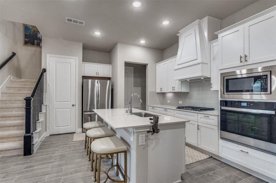 Kitchen with stainless steel appliances, white cabinets, a kitchen island with sink, backsplash, and custom exhaust hood