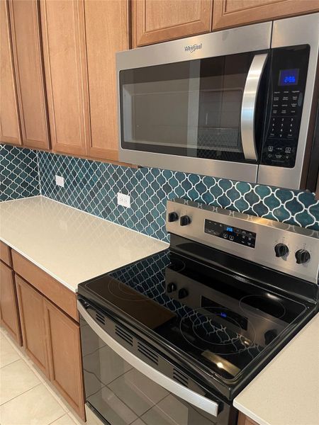 Kitchen featuring appliances with stainless steel finishes, backsplash, light tile patterned floors, light stone countertops, and brown cabinets Kitchen featuring appliances with stainless steel finishes, backsplash, light tile patterned floors, light stone countertops, and brown cabinets