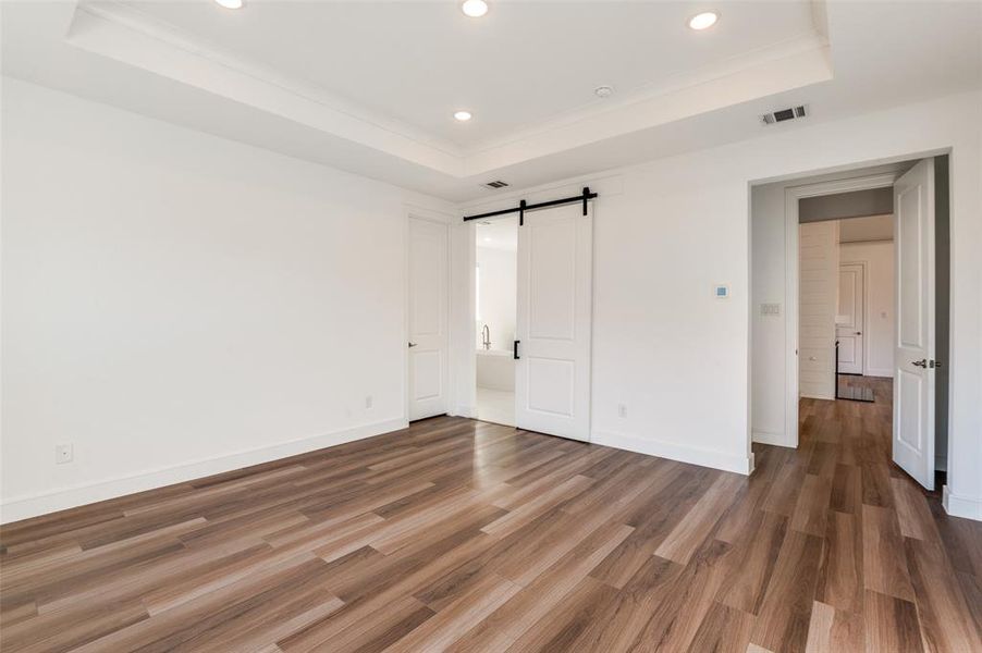 Primary bedroom with a tray ceiling, a barn door, wood finished floors, and recessed lighting Primary bedroom with a tray ceiling, a barn door, wood finished floors, and recessed lighting