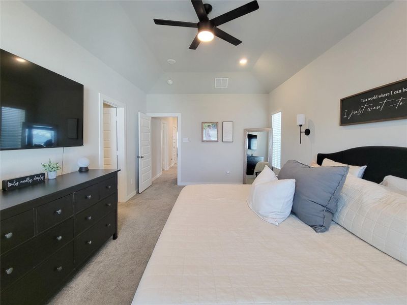This Primary bedroom features a spacious design with a neutral color palette and carpeted floor. It includes a large bed with multiple pillows, a sleek black ceiling fan, and a wall-mounted TV. The room is well-lit with natural light from windows.
