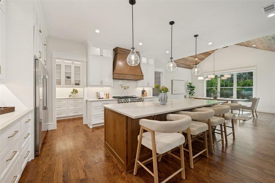 Expansive kitchen featuring wood-finish flooring, a large central island with seating, a wood-paneled vent hood, white cabinetry, and stainless steel appliances