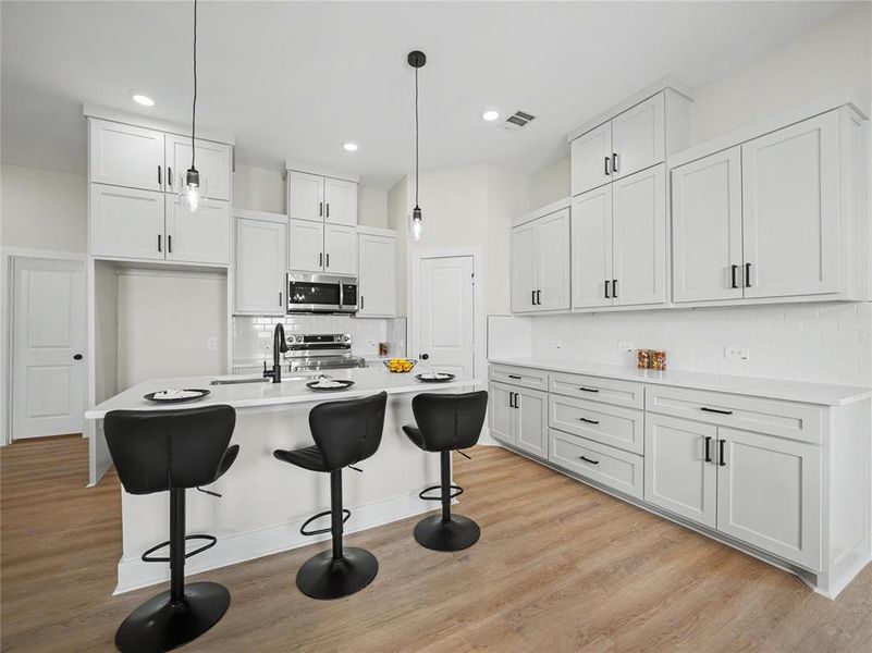 Contemporary kitchen featuring white shaker cabinetry, an island with countertop seating, stainless steel appliances, and wood-finish flooring Contemporary kitchen featuring white shaker cabinetry, an island with countertop seating, stainless steel appliances, and wood-finish flooring