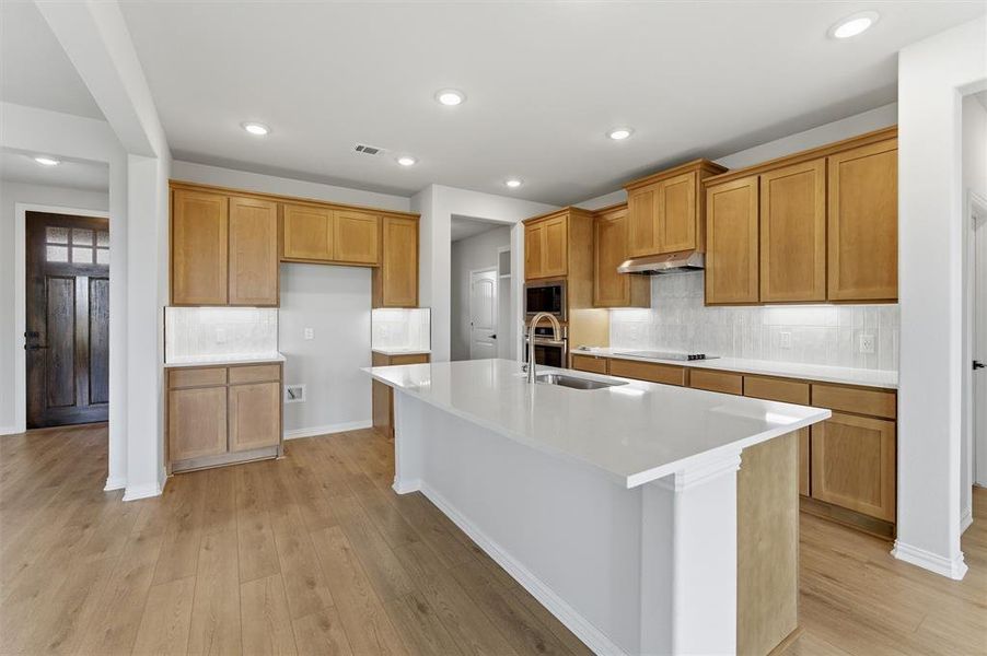 Kitchen with a kitchen island with sink, backsplash, light wood-type flooring, wood finish cabinetry, and recessed lighting