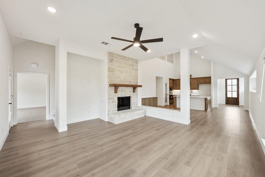 Open living room with stone fireplace, ceiling fan, hardwood floors, and open kitchen view beyond Open living room with stone fireplace, ceiling fan, hardwood floors, and open kitchen view beyond
