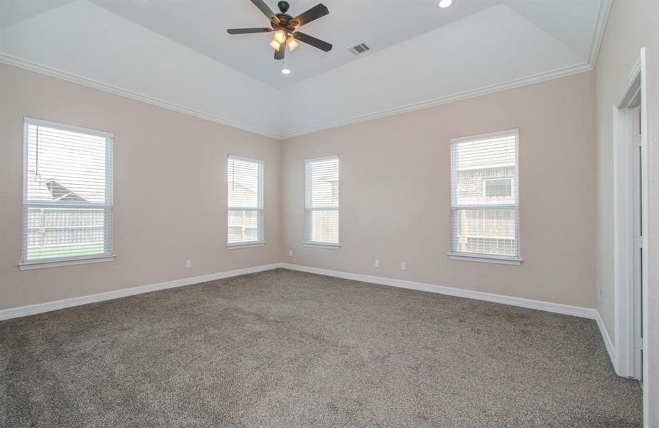 This is a spacious, well-lit room with neutral walls, plush carpet flooring, and a vaulted ceiling with a modern ceiling fan. It features three large windows that allow for ample natural light.