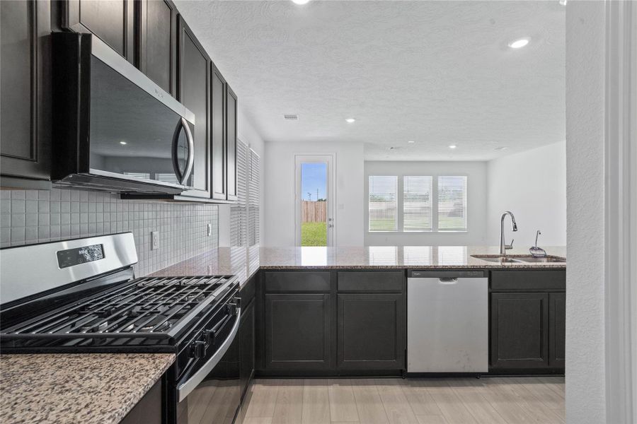 This spacious kitchen boasts high ceilings, Woodmont Belmont cabinets in an Espresso stain, Granite Caramel Cream countertops, stainless steel Whirlpool appliances, a modern Daltile backsplash, recessed lighting, an extended breakfast bar countertop, and a pantry—all overlooking the expansive Great Room.