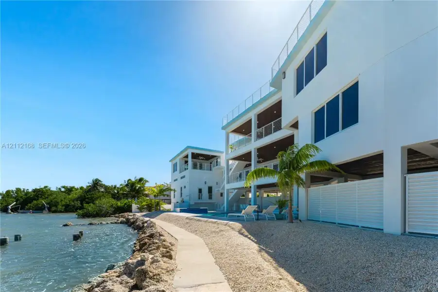 Multi-level waterfront exterior featuring white stucco siding, glass-panel railings, covered balconies, an inground swimming pool, and stone-lined shoreline Multi-level waterfront exterior featuring white stucco siding, glass-panel railings, covered balconies, an inground swimming pool, and stone-lined shoreline