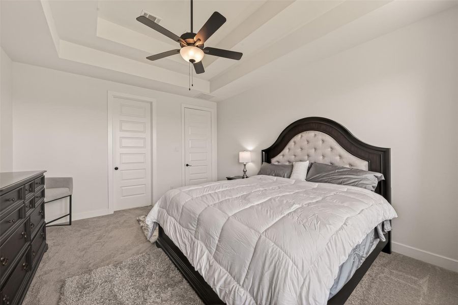 This spacious owners' retreat features a plush bed with an elegant headboard, a ceiling fan, and soft carpet flooring. Dual white doors and a dark dresser add style and function. Ideal for relaxation.