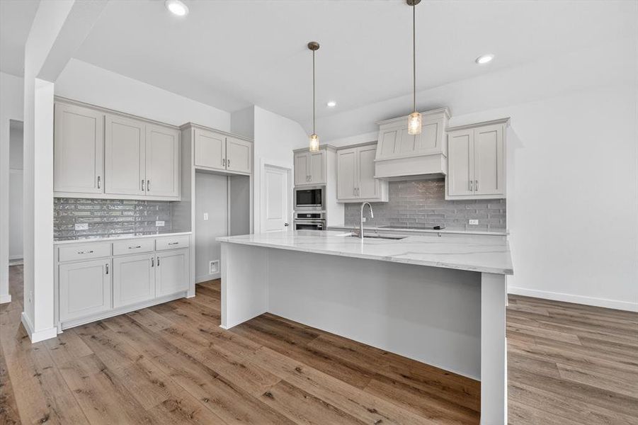 Gourmet kitchen featuring a large central island with a sink and pendant lighting, light-toned cabinetry, subway tile backsplash, and wood-finish flooring