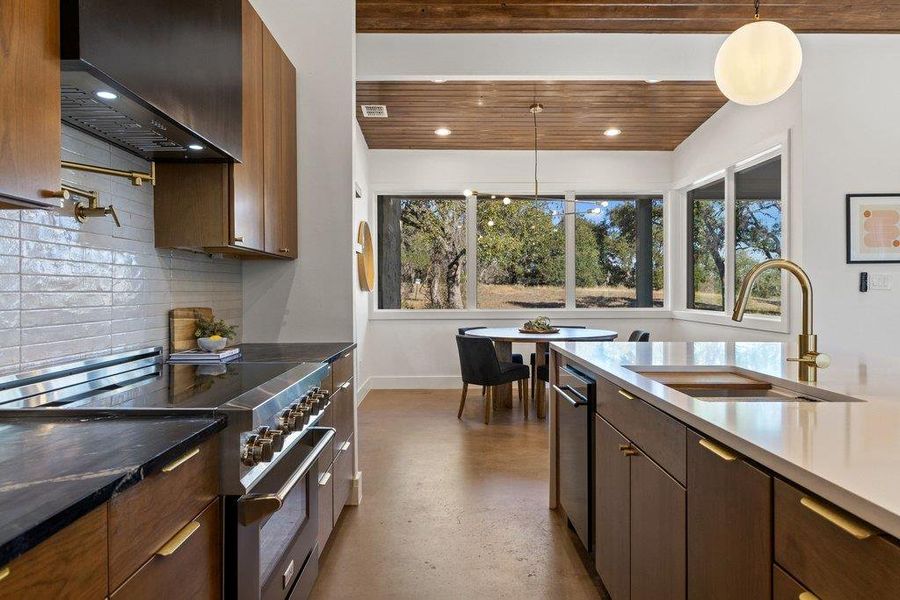 Kitchen featuring stainless steel stove, wall chimney range hood, hanging light fixtures, finished concrete floors, and dark stone countertops
