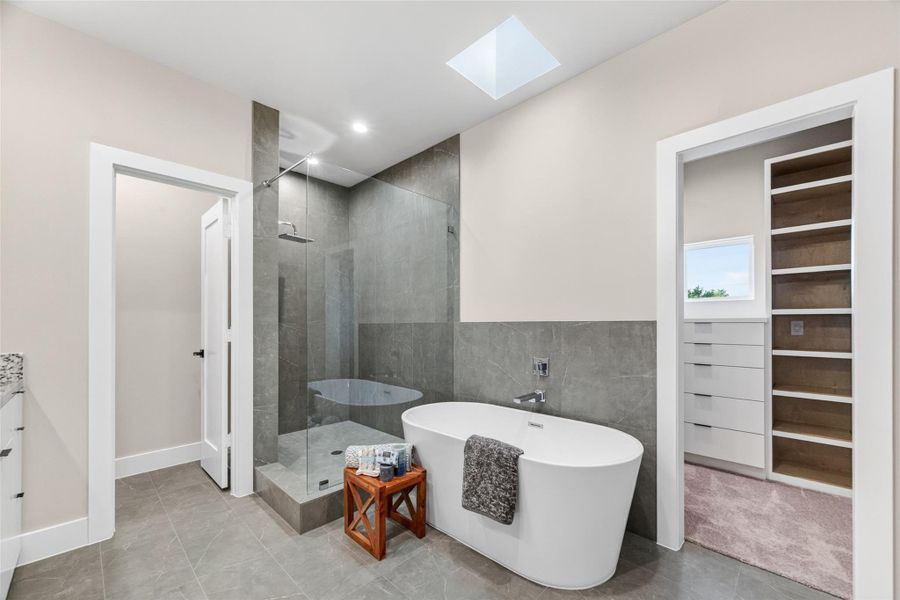 The primary bath delivers a spa-like experience: a freestanding soaking tub, an oversized walk-in shower finished in large-format tile with a glass panel and rain showerhead, and a skylight above that floods the space with natural light. The custom walk-in closet is visible through the doorway beyond.
