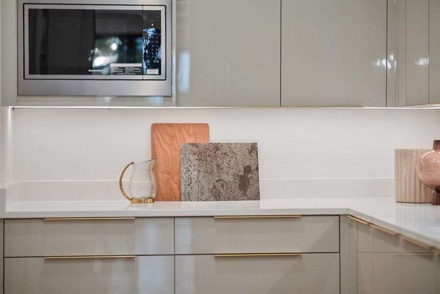 Sleek kitchen featuring a built-in stainless steel microwave, glossy light gray cabinetry, under-cabinet lighting, and white countertops