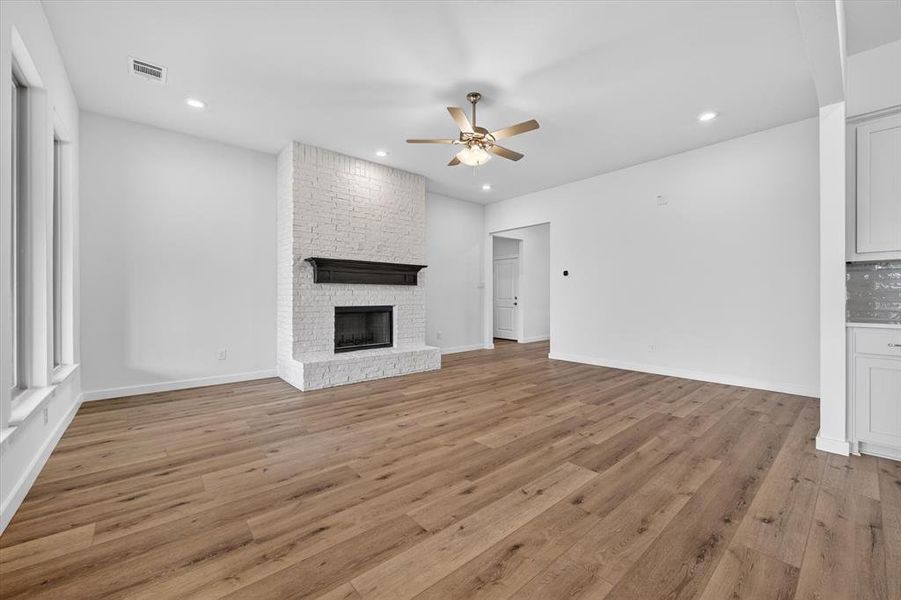 Spacious room featuring wood-finish flooring, a white brick fireplace with a dark mantel, recessed lighting, a ceiling fan, and large windows