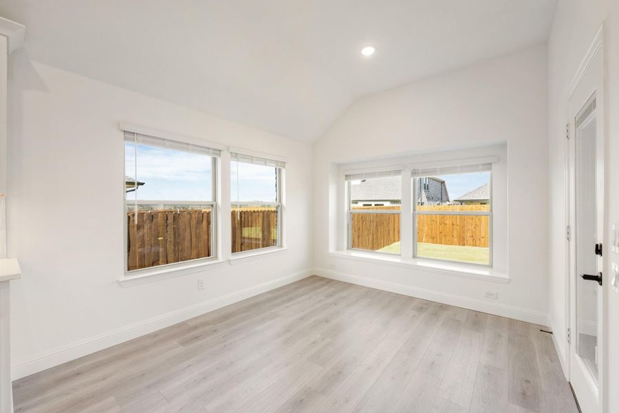Empty room with vaulted ceiling, light wood floors, and multiple windows overlooking a wood fence backyard Empty room with vaulted ceiling, light wood floors, and multiple windows overlooking a wood fence backyard