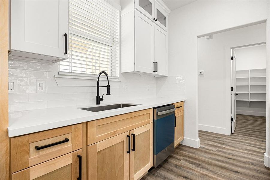 This modern kitchen features light wood cabinets, white countertops, and a sleek black faucet. A dishwasher is installed, and there's a view into a pantry with ample shelving. The room is brightened by a window with blinds and complemented by stylish wood flooring.