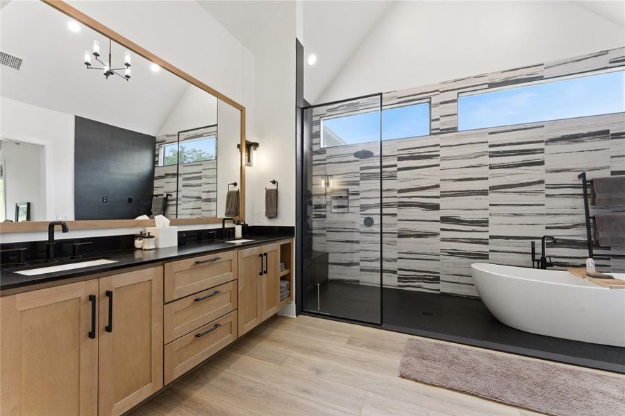 Contemporary bathroom featuring a freestanding soaking tub, a spacious walk-in shower with glass enclosure, a dual vanity with light wood cabinetry and dark countertops, wood-finish flooring, and horizontal accent windows Contemporary bathroom featuring a freestanding soaking tub, a spacious walk-in shower with glass enclosure, a dual vanity with light wood cabinetry and dark countertops, wood-finish flooring, and horizontal accent windows