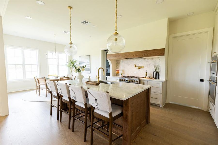 Gourmet kitchen featuring a large island with seating, a professional-grade range with a custom wood-beam hood, double wall ovens, marble-look countertops, and wood-finish flooring