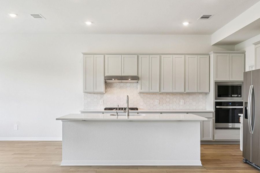 Kitchen with decorative backsplash, stainless steel appliances, light wood finished floors, a center island with sink, and light stone countertops Kitchen with decorative backsplash, stainless steel appliances, light wood finished floors, a center island with sink, and light stone countertops