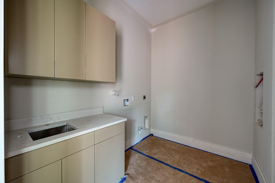 A utility room with neutral walls, featuring upper and lower cabinets with a countertop and sink
