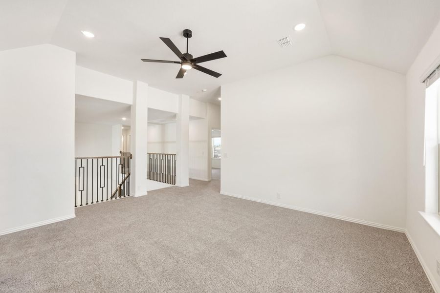 Open upstairs living area with carpet, vaulted ceiling, ceiling fan, and staircase railing nearby.