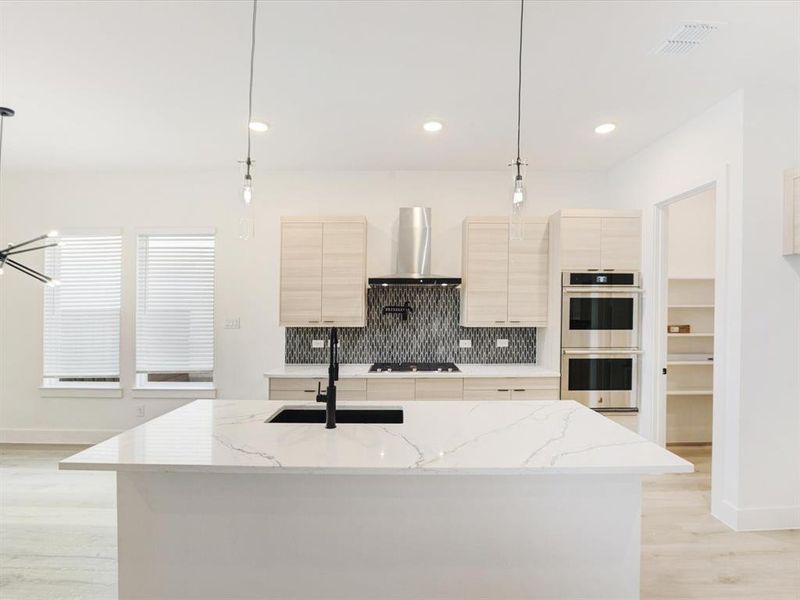 Kitchen with backsplash, light stone counters, hanging light fixtures, light brown cabinets, and light wood finished floors Kitchen with backsplash, light stone counters, hanging light fixtures, light brown cabinets, and light wood finished floors