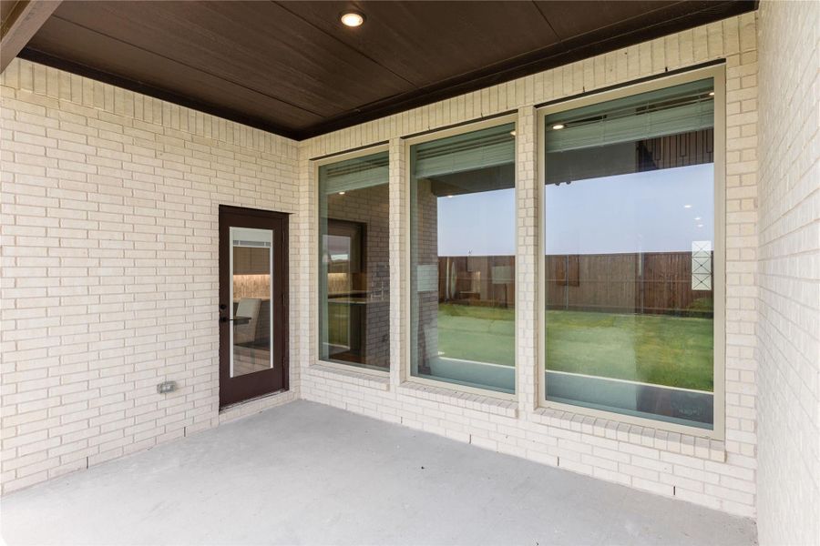 Covered patio with white brick exterior, large windows, and dark wood ceiling overlooking a fenced backyard