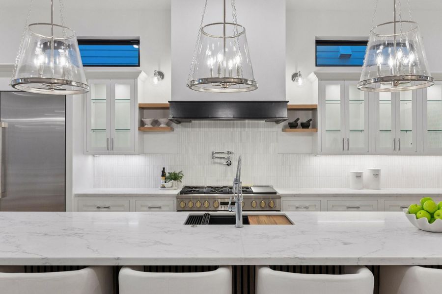 A closer view of this gorgeous kitchen showcases Slab Quartz Counter, 1 x 6 Honed Stacked Marble Backsplash, 33 Inch Undermount Single Basin Sink in Quartz Composite Finish, Pull-Down Faucet in Polished Chrome and Top Switch Disposal, 24" Pot Filler in a Polished Chrome Finish, White Oak Floating Shelves with 6" LED Sconces  in a Chrome Finish with Clear Shades, LED Under-Cabinet Lighting and Under-Cabinet PlugMold. Inset Cabinetry with Glass Fronted Doors and Shelves with Lighting on the Upper Cabinets.