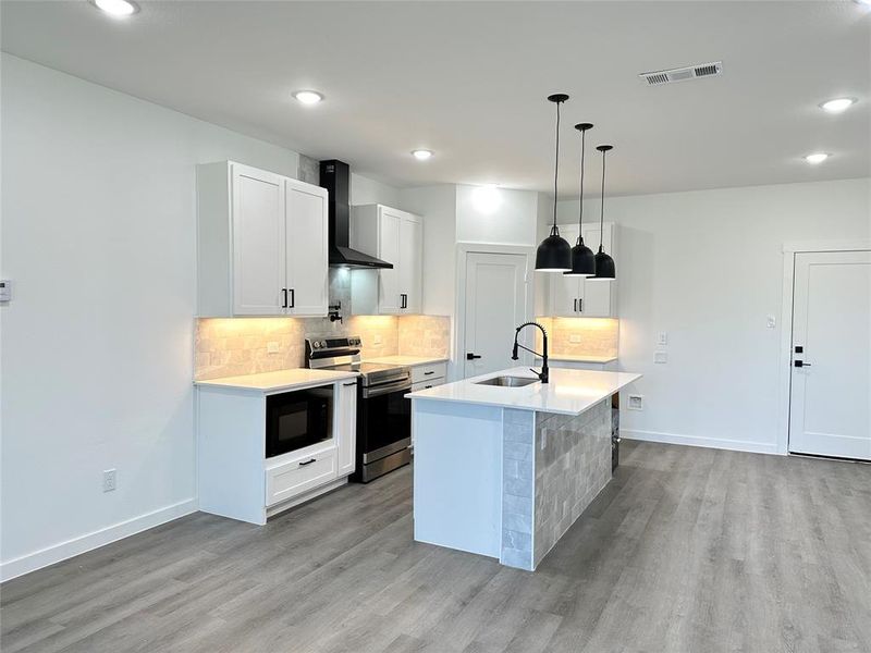 Kitchen featuring sink, wall chimney exhaust hood, stainless steel range with electric stovetop, an island with sink, and white cabinets