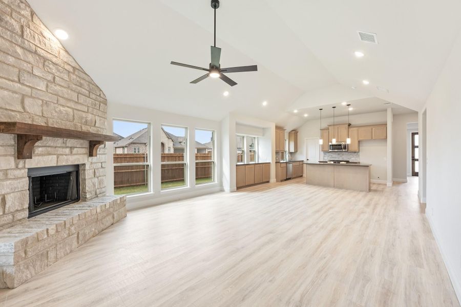 Open living area with stone fireplace, light wood floors, and open kitchen with island visible in background