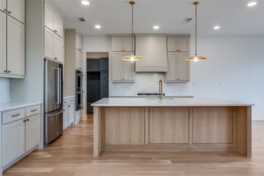 Kitchen with tasteful backsplash, hanging light fixtures, a kitchen island with sink, stainless steel appliances, and light hardwood / wood-style flooring